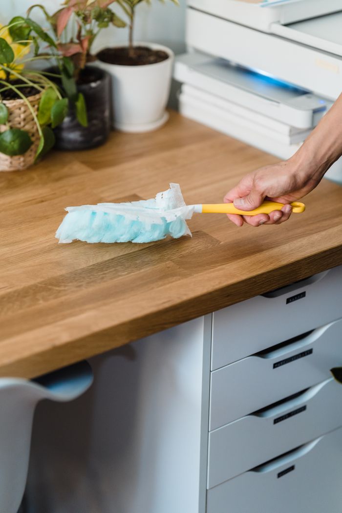 A hand with a feather duster cleaning a wooden desk, with plants and office supplies around it.
